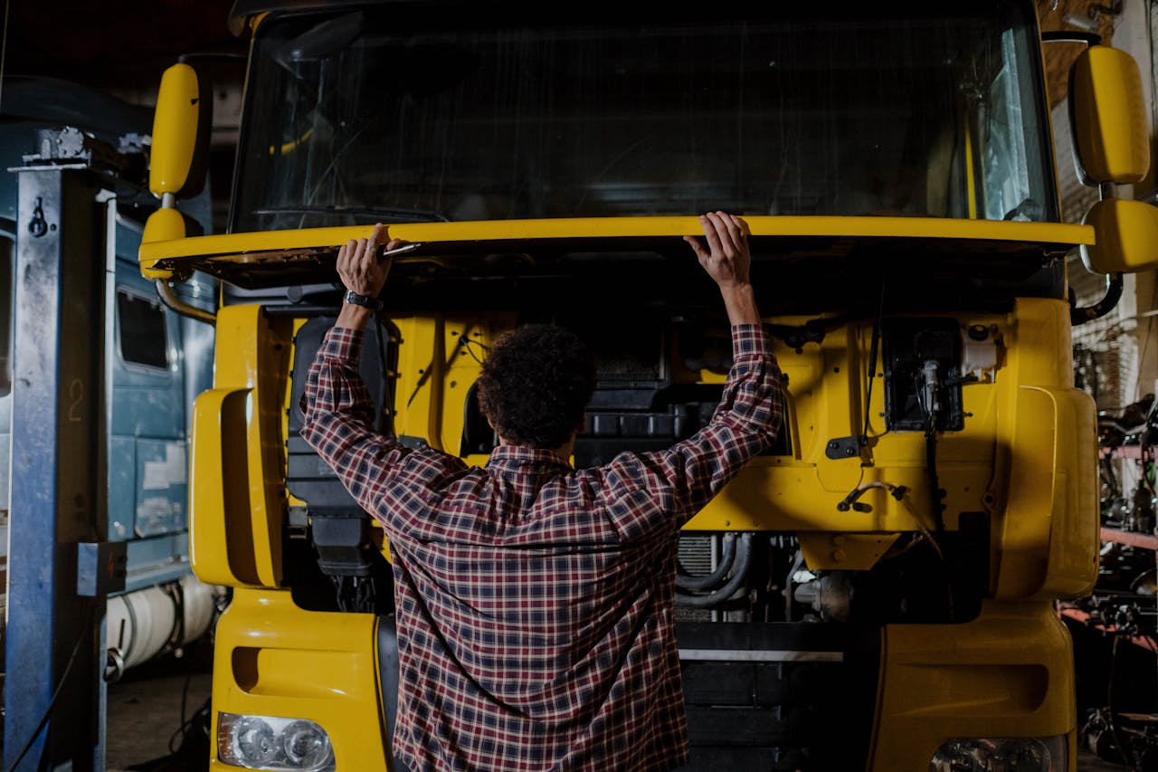 Rear view of a mechanic inspecting a yellow truck engine in a garage setting.